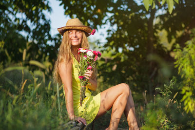 Side view of woman wearing hat holding flowers while sitting on land against trees