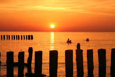 Silhouette people swimming in sea by wooden posts against sky during sunset
