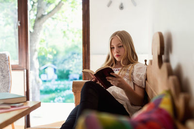 Teenage girl reading book while sitting on sofa at home