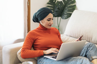 Muslim woman in casual clothes and headscarf browsing social media on cellphone while sitting with laptop on couch and listening to music on weekend day at home