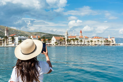 Rear view of young woman using mobile phone, taking photos of beautiful cityscape of trogir, croatia