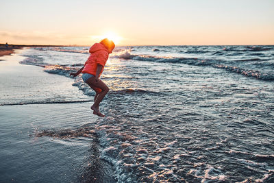 Playful little girl jumping over sea waves on a sand beach at sunset during summer vacation