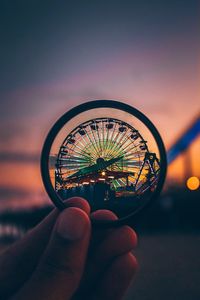 Cropped hand holding lens against ferris wheel during sunset