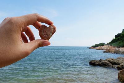 Cropped hand of woman holding heart shape pebble against sea and sky