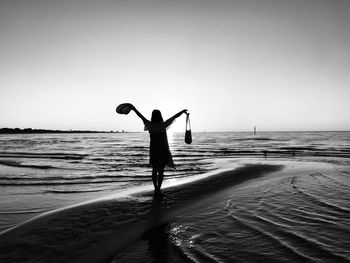 Silhouette woman holding bag and hat standing in beach against sky