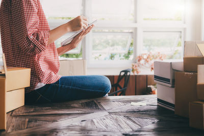 Midsection of woman holding digital tablet while sitting by cardboard boxes on table