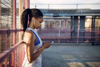 Side view of woman using phone while standing against fence