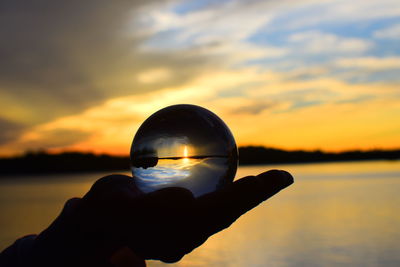 Reflection of hand holding crystal ball against sky during sunset