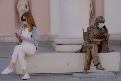 Woman wearing mask using mobile phone while sitting by statue in city