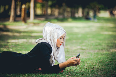 Side view of woman wearing hijab while using mobile phone on grassy field at park