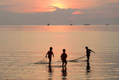 Silhouette children with fishing net standing in sea against sky during sunset