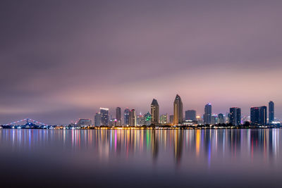 Illuminated city buildings by bay against sky