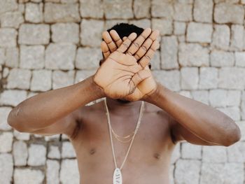 Shirtless man covering face with hands while standing against brick wall