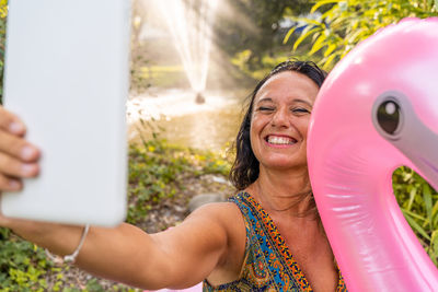 Beautiful smiling middle aged woman taking a selfie sitting on a pink flamingo shaped inflatable toy