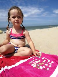 Cute girl in swimwear sitting on blanket at beach against blue sky during sunny day