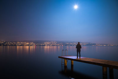 Man standing on pier over lake against illuminated city at night
