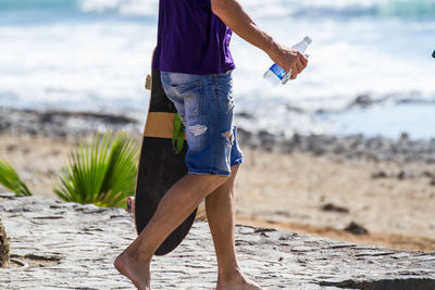 Midsection of man with bottle and skateboard walking at beach