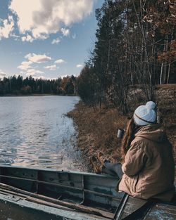 Rear view of woman sitting by river against sky during winter