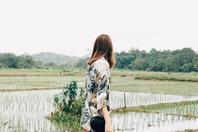 Rear view of person standing on field by lake against clear sky