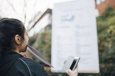 Female messenger carrying document in mouth and holding smart phone looking at billboard