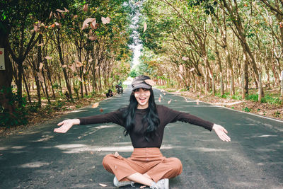 Portrait of smiling young woman throwing leaves while sitting on road amidst trees in forest