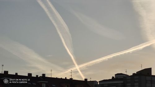 Low angle view of vapor trails over silhouette buildings against sky