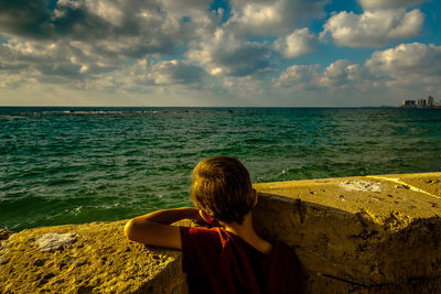Rear view of boy standing by retaining wall in front of sea