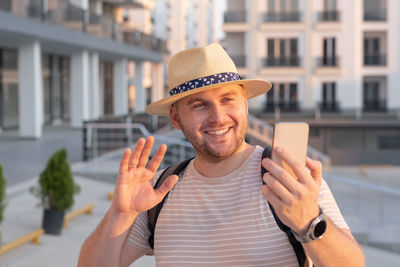 Handsome blond bearded smiling man,happy tourist,wearing sunhat,backpack,video call,using smartphone