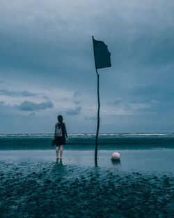 Rear view of woman with backpack standing at beach against cloudy sky