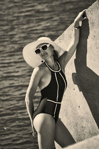 Portrait of a woman in a swimsuit, hat and sunglasses in summer on the riverbank by a concrete wall