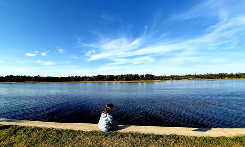 Woman sitting by lake against blue sky