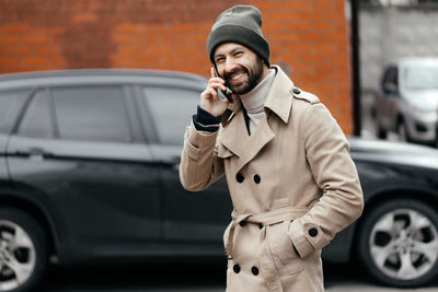 Close-up of a young, smiling, happy, laughing hipster with a beard in a raincoat and hat,