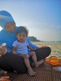 Portrait of girl with mother sitting at beach against blue sky