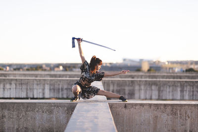 Young female athlete practicing sword on structure against clear sky during sunset