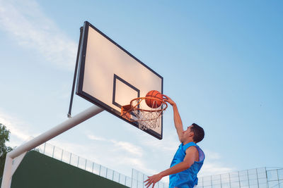 Full length of man jumping with ball by basketball hoop against sky