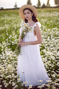 Beautiful young brunette woman in a hat and a white dress standing on a chamomile field at sunset