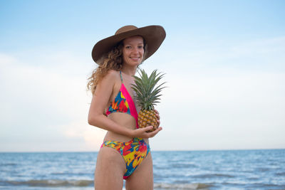 Portrait of young woman wearing hat and holding pineapple against sky