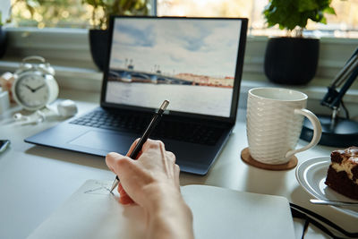 Designer draws sketch on paper, sits at desk with laptop in front of window