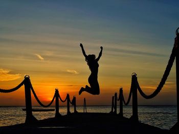 Silhouette woman jumping over pier at beach against sky during sunset