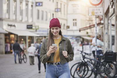Smiling young woman using smart phone while listening music in city