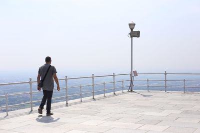 Rear view of man standing on footpath by sea against clear sky