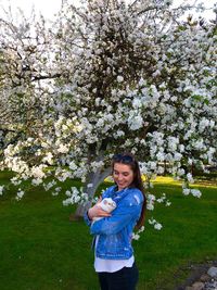 Smiling woman holding guinea pig while standing by flowering tree 