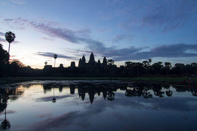 Silhouette buildings by lake against sky at sunset