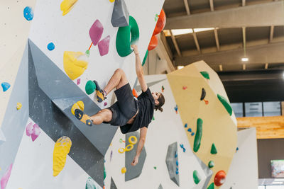 Full body side view of sportive male alpinist climbing wall with colorful grips during intense training in light modern gym