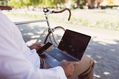 Midsection of businessman using laptop and smart phone while sitting on park bench
