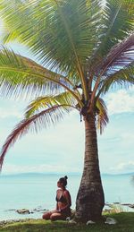 Woman sitting on palm tree by sea against sky