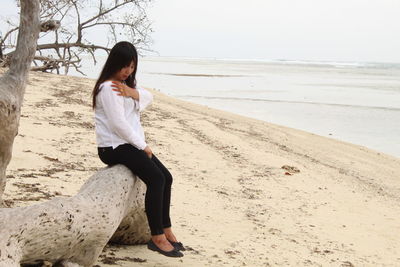 Full length of young woman sitting on driftwood at beach