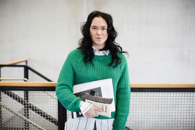 Portrait of smiling female student with books standing at railing against wall in university