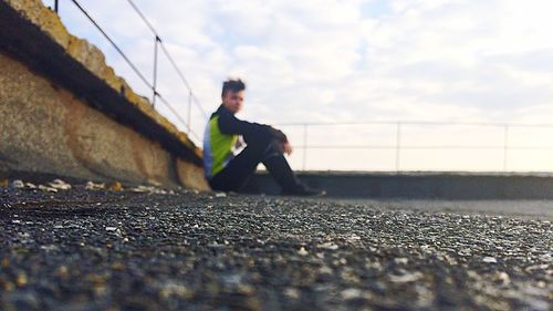Surface level view of man sitting on footpath by retaining wall against sky