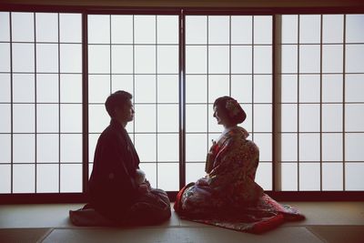 Smiling bride and groom sitting face to face against window during wedding